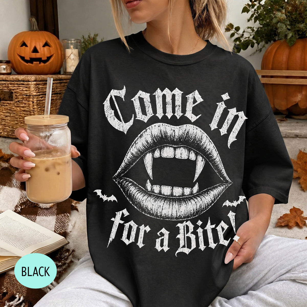 A woman wearing a black t-shirt with a graphic design of a pair of open-mouthed fangs and the text "Come in for a bite" is sitting on a couch with a pumpkin and a book nearby.