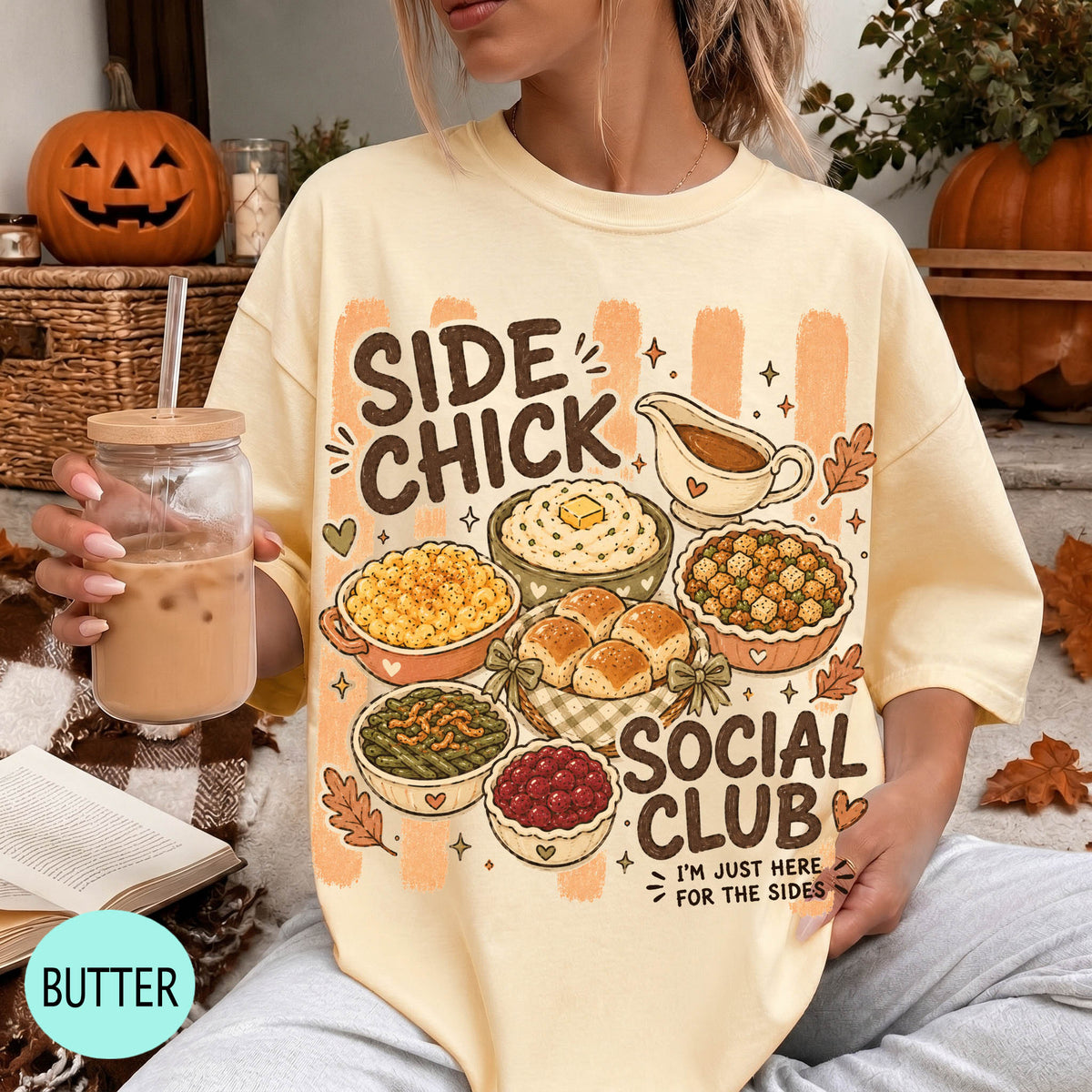 A person wearing a yellow t-shirt with a graphic design of various fall dishes and a caption that reads "Side Chick Social Club" is holding a drink and surrounded by fall decorations.