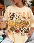 A person wearing a yellow t-shirt with a graphic design of various fall dishes and a caption that reads "Side Chick Social Club" is holding a drink and surrounded by fall decorations.