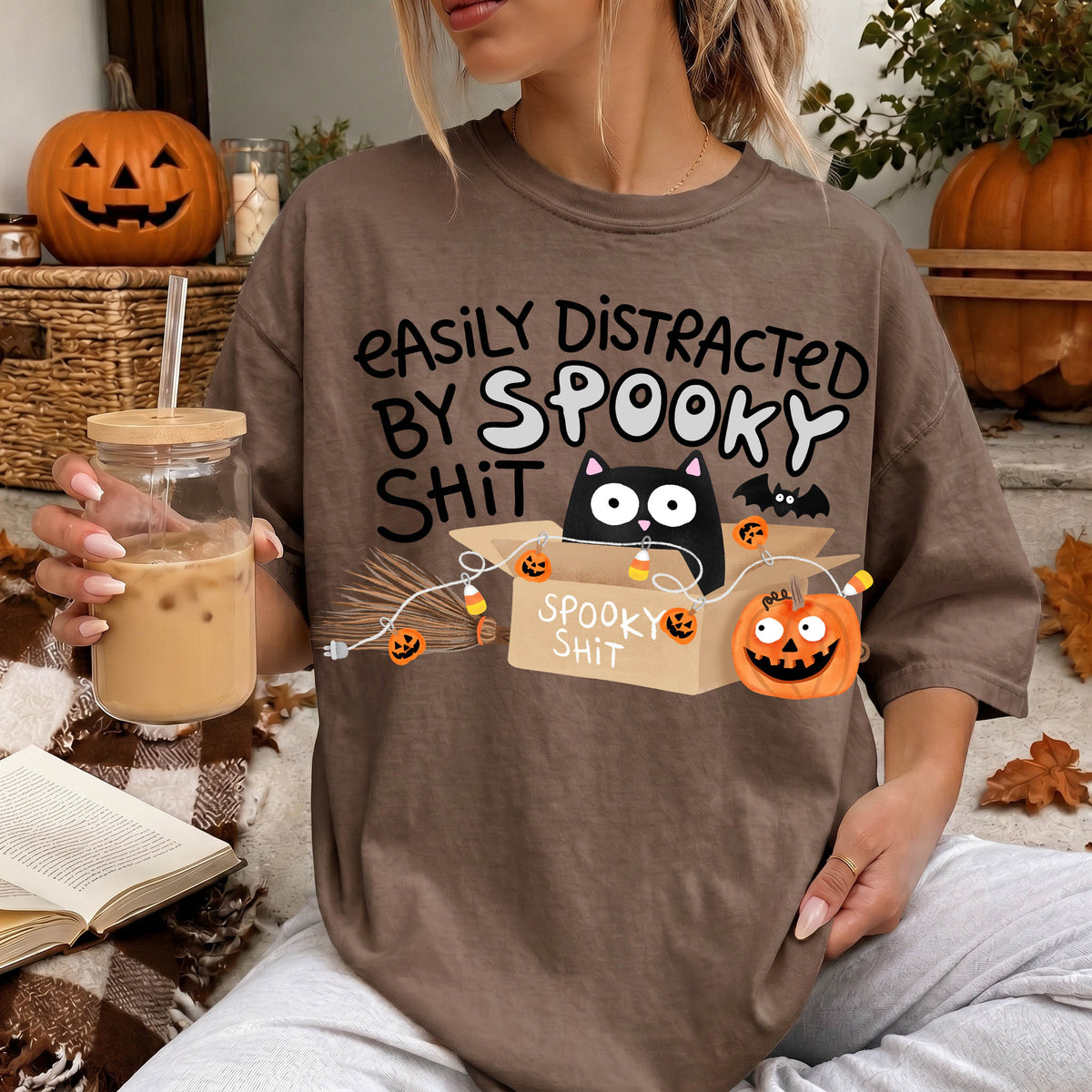 A person wearing a t-shirt with a humorous Halloween-themed graphic, including a cat, pumpkin, and text that says "Easily distracted by Spooky Shit".