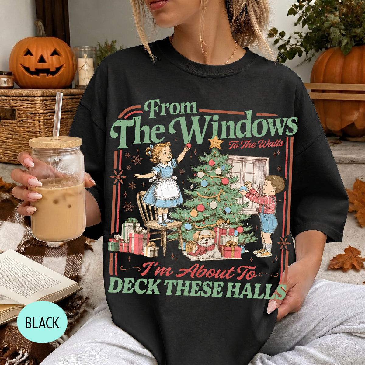 A woman wearing a black t-shirt with a graphic design depicting a Christmas tree and two children decorating it. She is holding a drink in her hand.