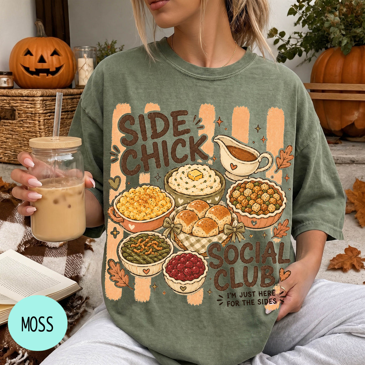 A woman wearing a green t-shirt with a graphic design of various Thanksgiving dishes and a caption that reads "SIDE CHICK SOCIAL CLUB I'M JUST HERE FOR THE SIDES". She is holding a drink in a glass with a straw.