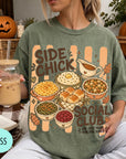A woman wearing a green t-shirt with a graphic design of various Thanksgiving dishes and a caption that reads "SIDE CHICK SOCIAL CLUB I'M JUST HERE FOR THE SIDES". She is holding a drink in a glass with a straw.