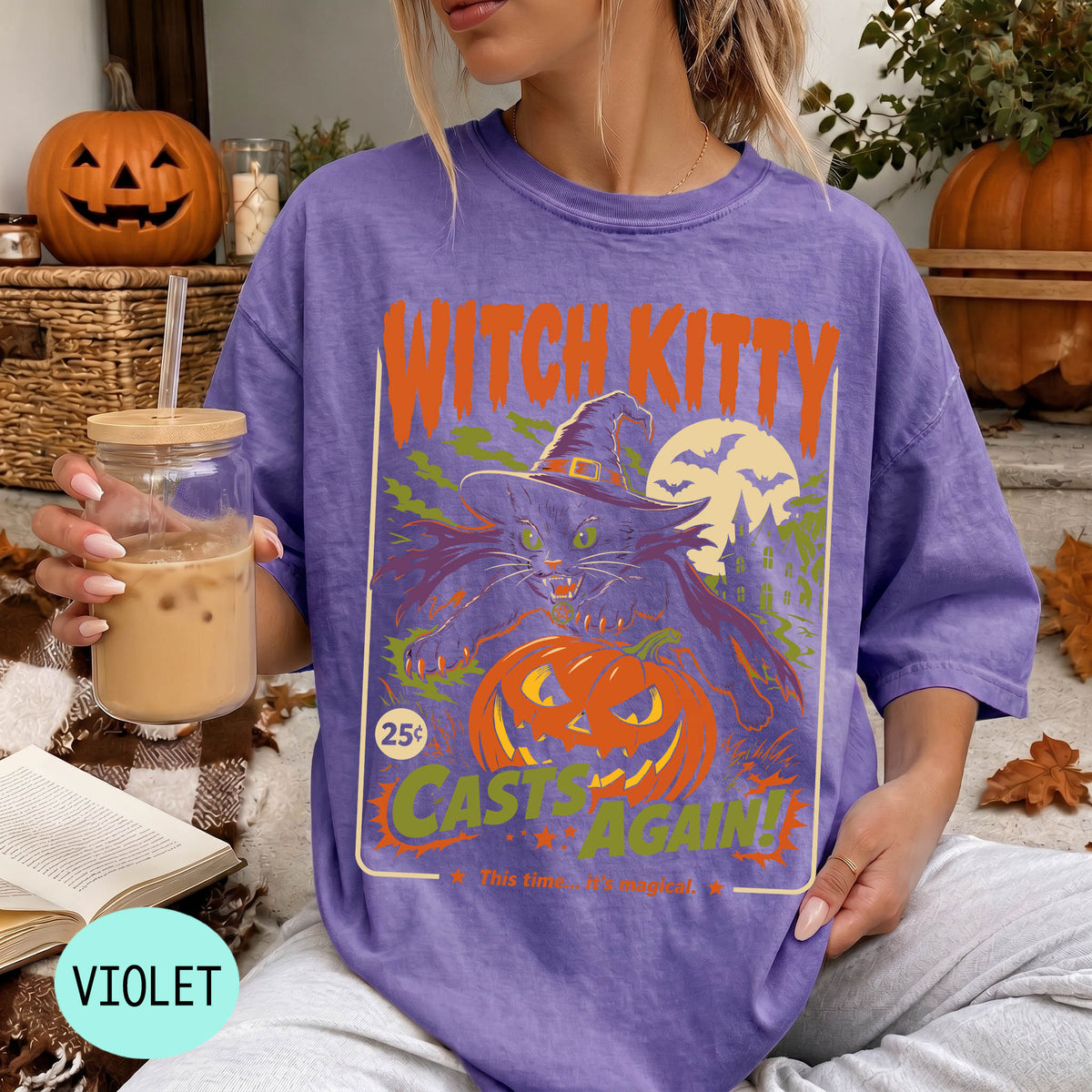 A woman wearing a purple t-shirt with a Halloween-themed graphic, holding a drink and surrounded by pumpkins and other fall decorations.
