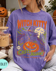 A woman wearing a purple t-shirt with a Halloween-themed graphic, holding a drink and surrounded by pumpkins and other fall decorations.