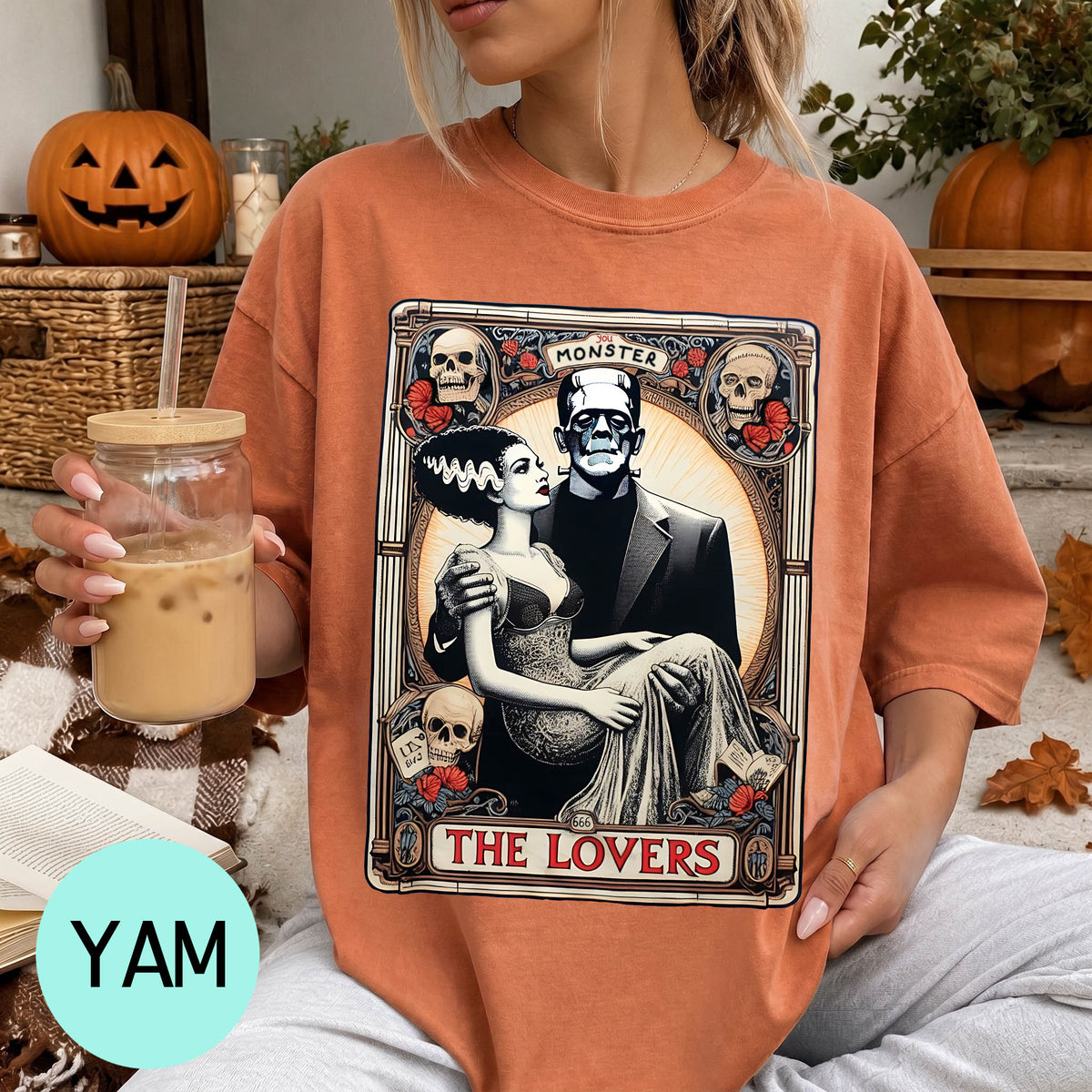 A woman wearing a t-shirt with a graphic design featuring a couple and the text "MONSTER THE LOVERS". She is holding a drink in a glass and there are Halloween decorations visible in the background.