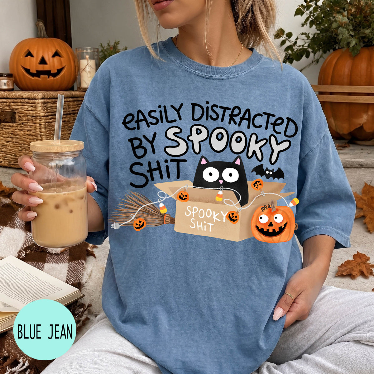 A woman wearing a blue t-shirt with a humorous Halloween-themed graphic, including a cat, pumpkin, and text that says "Easily Distracted by Spooky Shit". She is holding a drink in a glass with a straw.