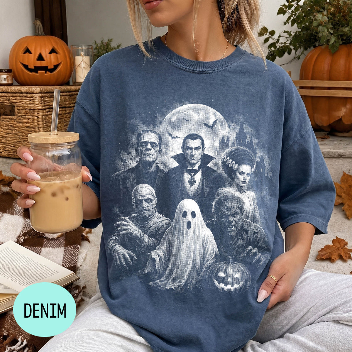 A woman wearing a blue t-shirt with a graphic design of a group of Halloween-themed characters, including Dracula, Frankenstein, Frankenstein's Bride, werewolf, and ghost, along with a pumpkin and other Halloween decorations. She is holding a drink in a glass with a straw.