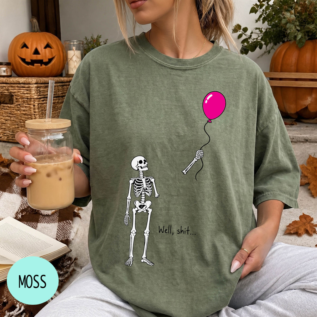 A woman wearing a green t-shirt with a skeleton holding a pink balloon and the text "Well, shit..." printed on it.