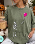 A woman wearing a green t-shirt with a skeleton holding a pink balloon and the text "Well, shit..." printed on it.