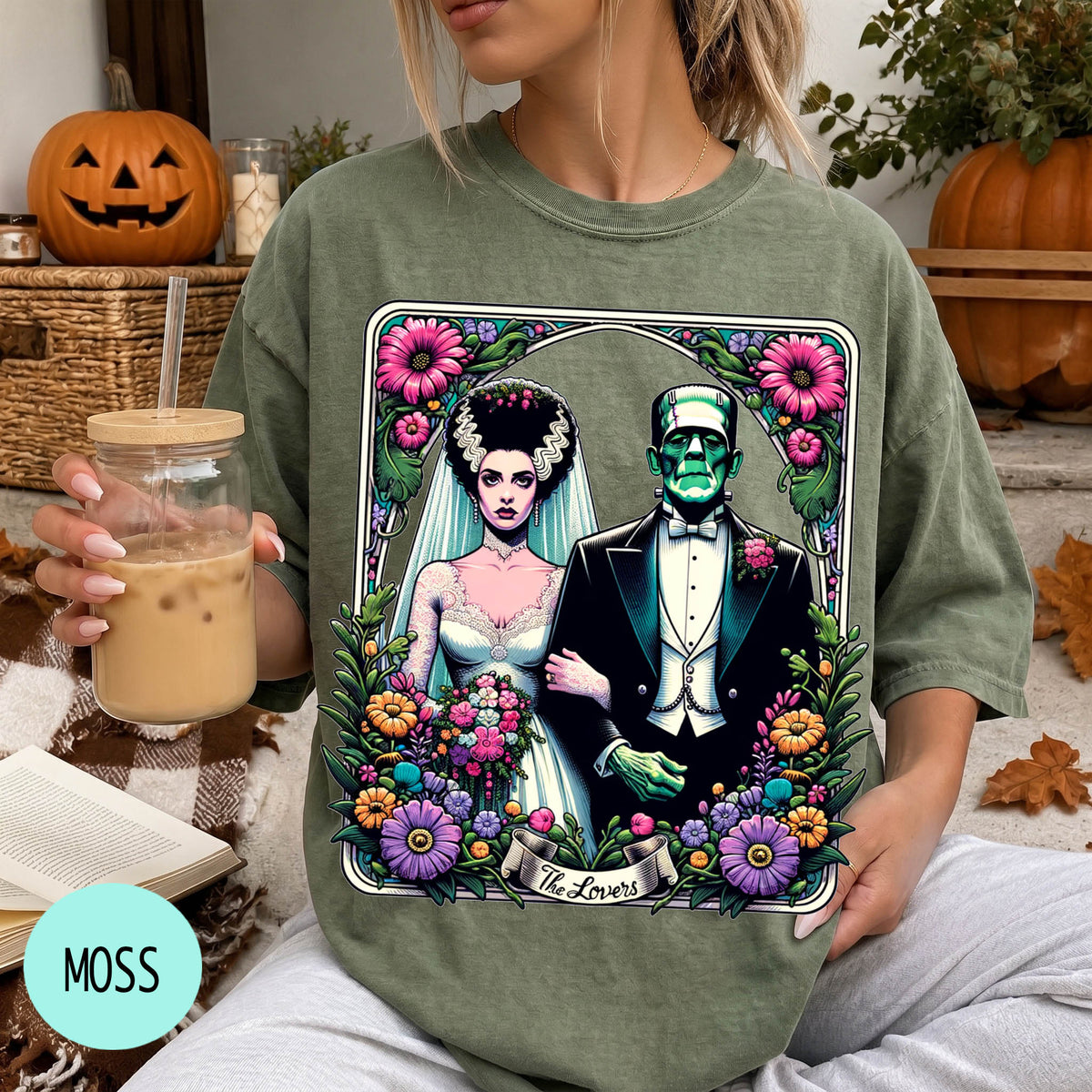 A woman wearing a green t-shirt with a graphic design of a bride and groom surrounded by flowers and a banner that says "The Lovers". She is holding a drink in a glass.