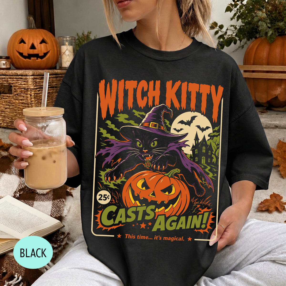 A woman wearing a black t-shirt with a Halloween-themed graphic design, including a witch, pumpkin, and bats. She is holding a drink in a glass with a straw.