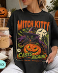 A woman wearing a black t-shirt with a Halloween-themed graphic design, including a witch, pumpkin, and bats. She is holding a drink in a glass with a straw.