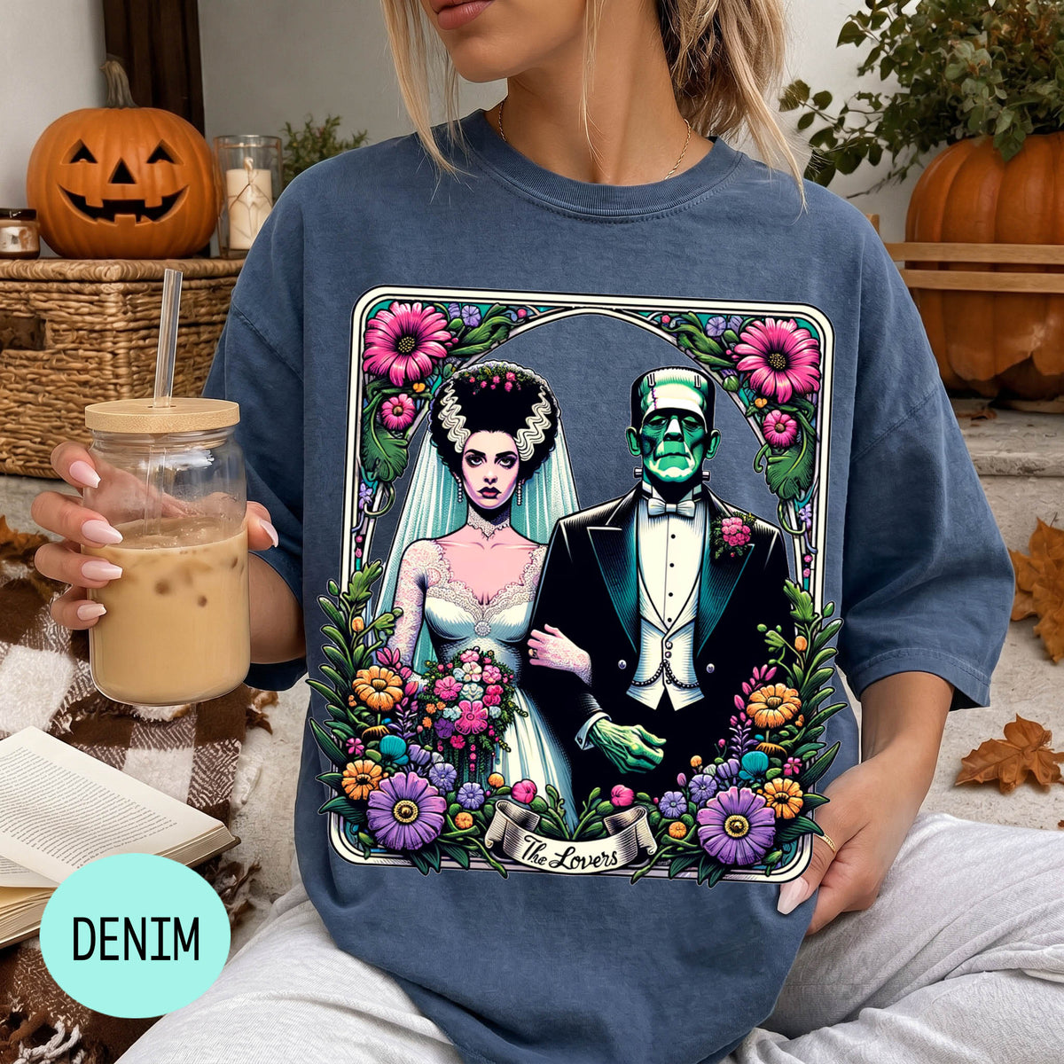 A woman wearing a blue t-shirt with a graphic design of a bride and a Frankenstein monster, holding a drink and surrounded by Halloween decorations.