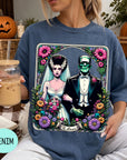 A woman wearing a blue t-shirt with a graphic design of a bride and a Frankenstein monster, holding a drink and surrounded by Halloween decorations.