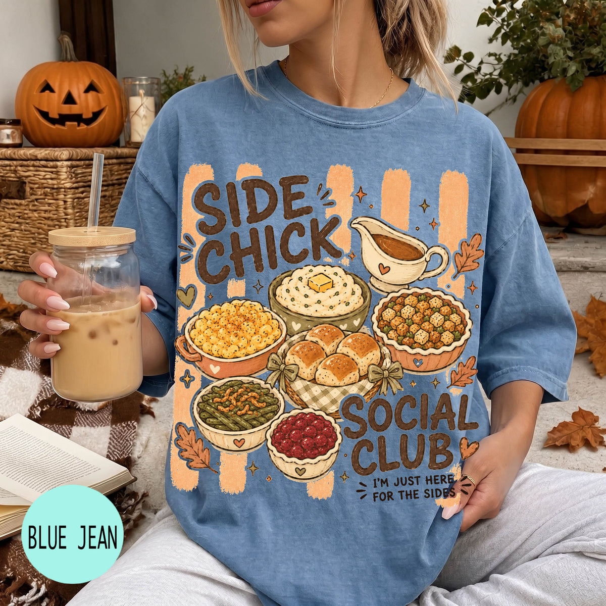 A woman wearing a blue t-shirt with a graphic design of various Thanksgiving dishes and the text "SIDE CHICK SOCIAL CLUB I'M JUST HERE FOR THE SIDES" is holding a drink and surrounded by fall decorations.