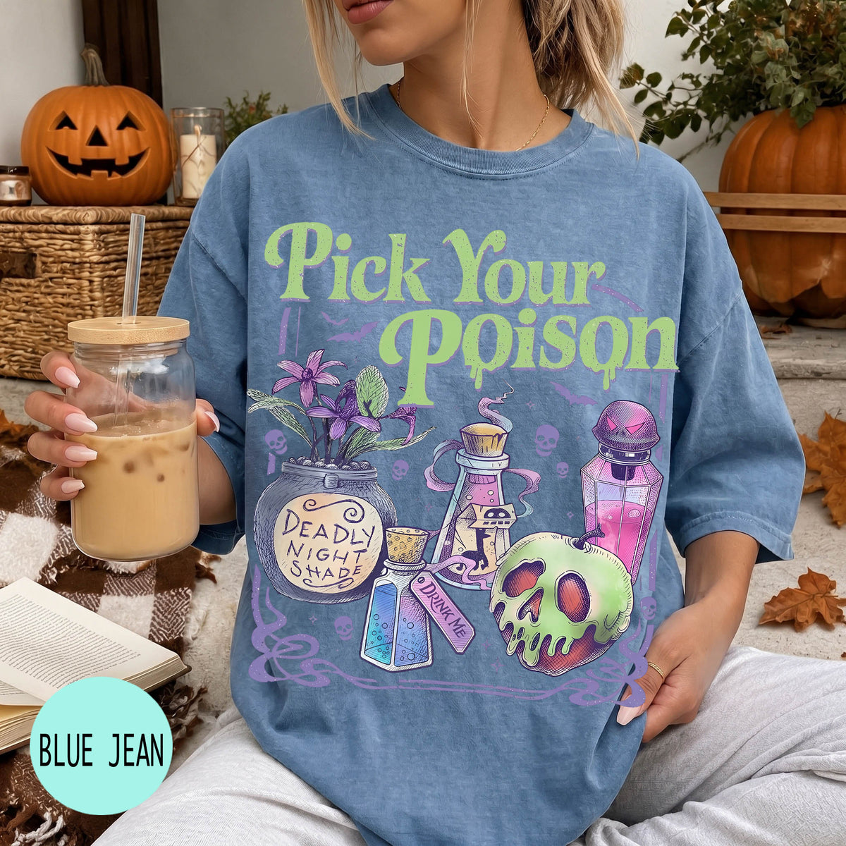 A woman wearing a blue t-shirt with a graphic design featuring various Halloween-themed items, such as a skull, a vase, and a bottle, along with the text "Pick Your Poison". She is holding a drink in a glass with a straw.