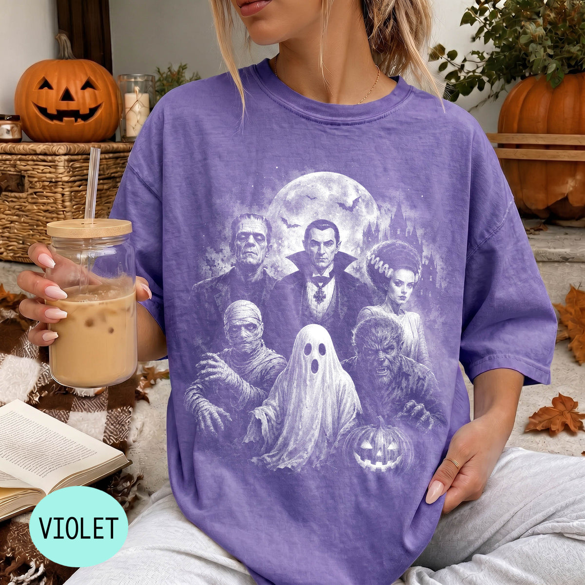 A person wearing a purple t-shirt with a Halloween-themed graphic of Dracula, Frankenstein, and a ghost, holding a drink and sitting on a blanket with a pumpkin and other Halloween decorations in the background.