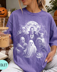 A person wearing a purple t-shirt with a Halloween-themed graphic of Dracula, Frankenstein, and a ghost, holding a drink and sitting on a blanket with a pumpkin and other Halloween decorations in the background.