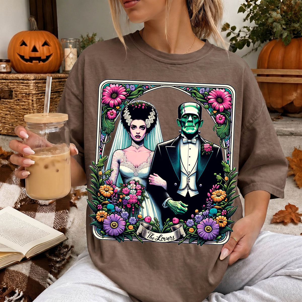 A woman is wearing a t-shirt with a graphic design featuring a bride and a Frankenstein monster, surrounded by flowers and a banner that says "The Lovers". She is holding a drink in a glass with a straw.