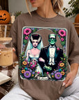 A woman is wearing a t-shirt with a graphic design featuring a bride and a Frankenstein monster, surrounded by flowers and a banner that says "The Lovers". She is holding a drink in a glass with a straw.