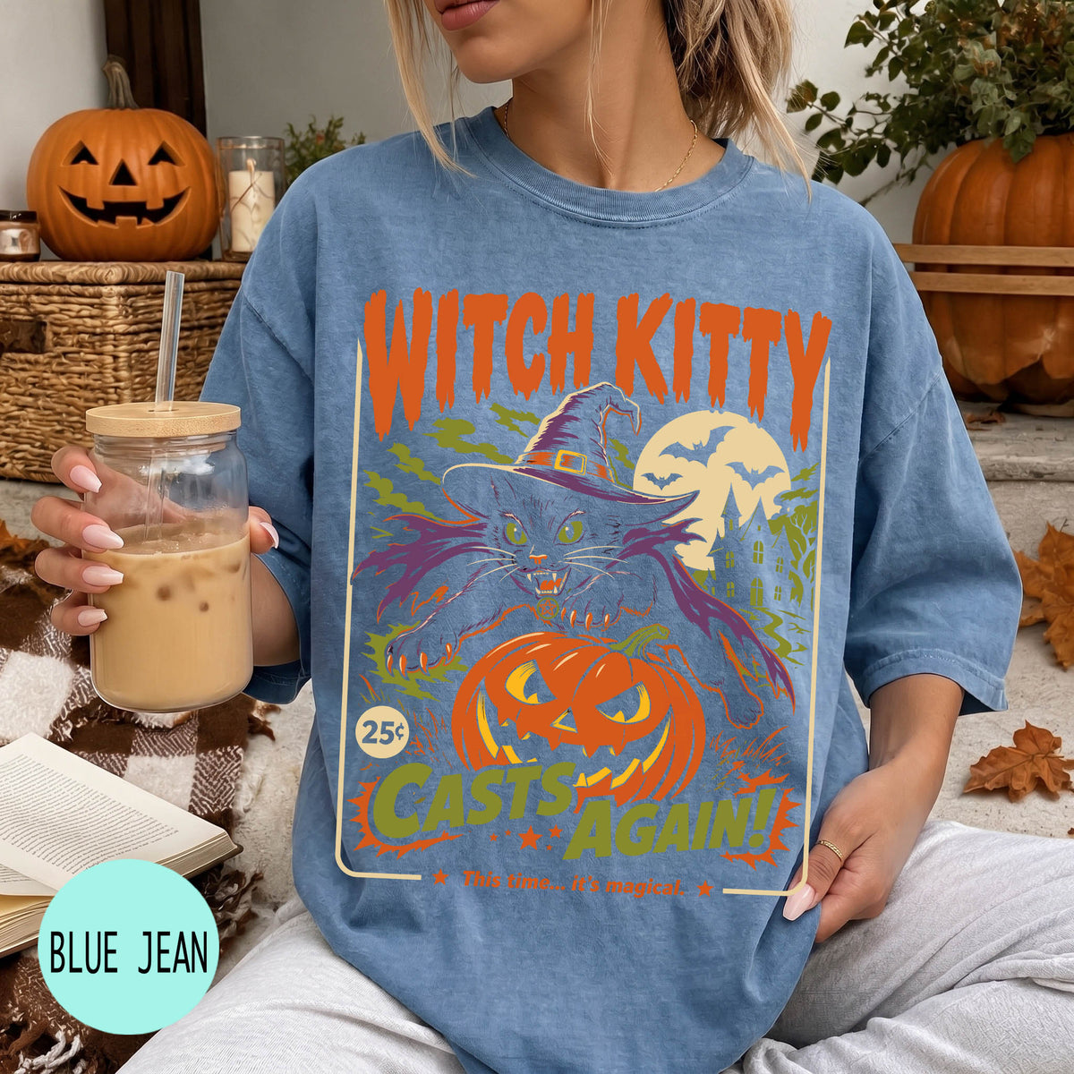 A woman wearing a blue t-shirt with a Halloween-themed graphic, holding a drink and surrounded by pumpkins and other fall decorations.