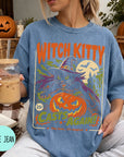 A woman wearing a blue t-shirt with a Halloween-themed graphic, holding a drink and surrounded by pumpkins and other fall decorations.