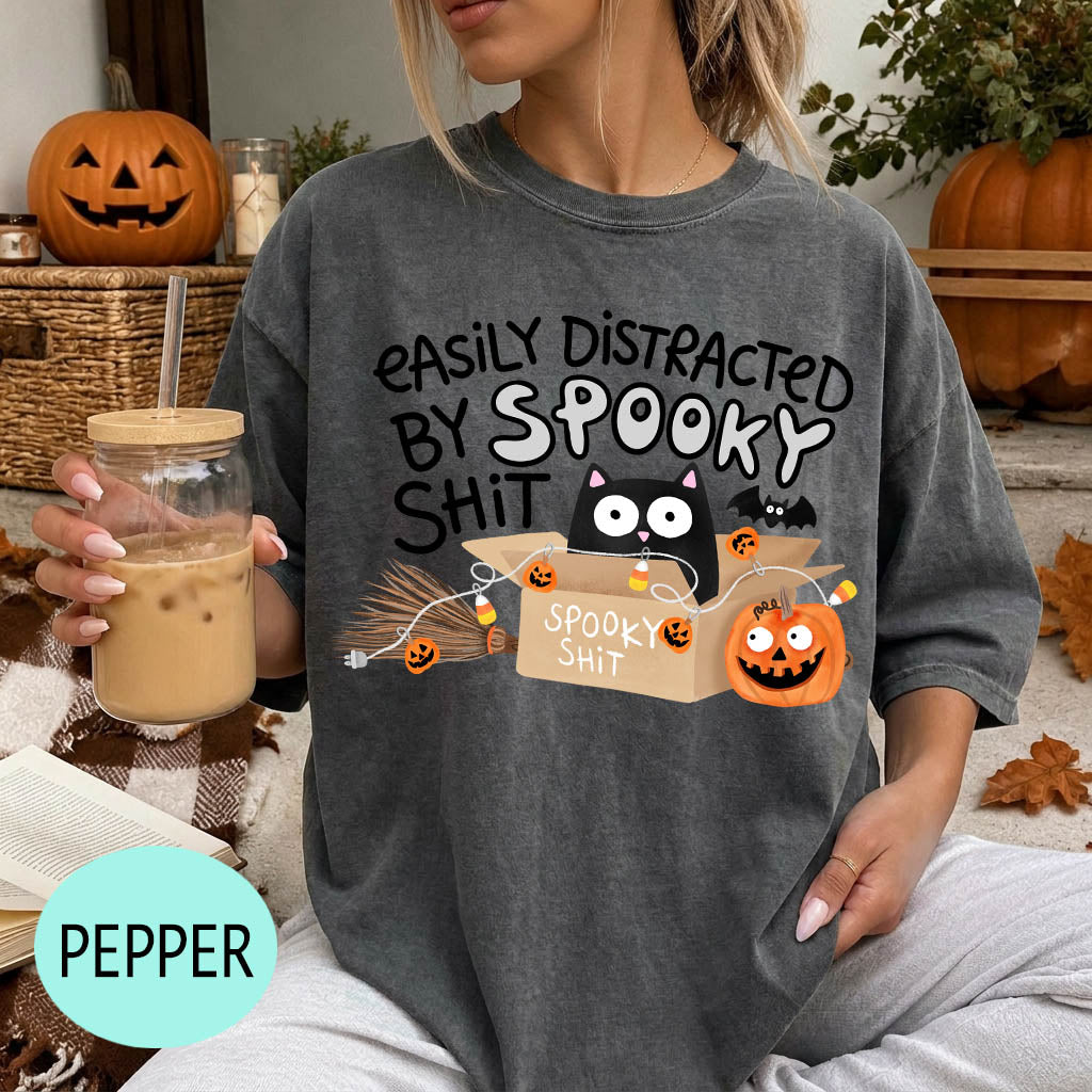 A woman wearing a gray t-shirt with a Halloween-themed graphic and text, holding a drink in a glass.