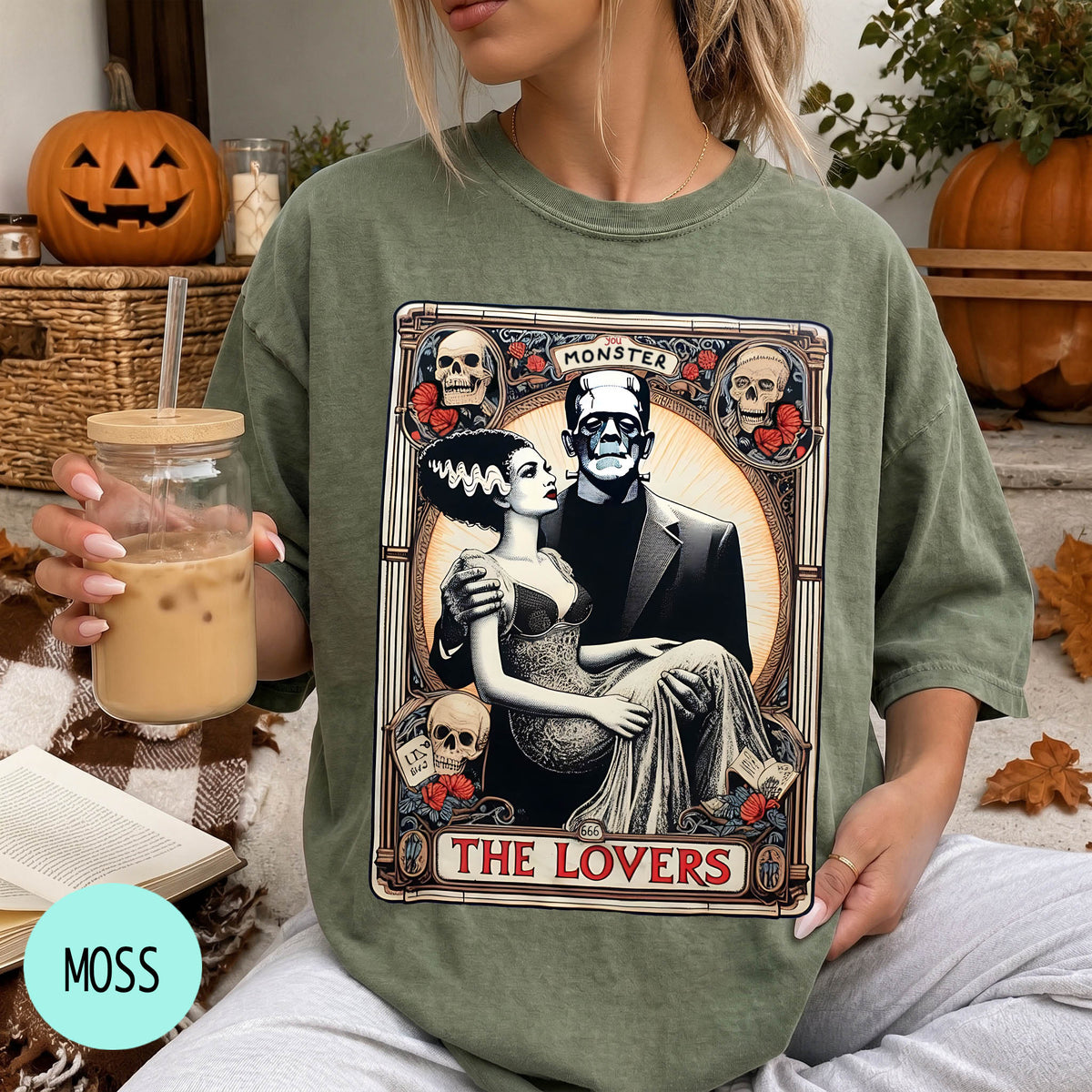 A woman wearing a green t-shirt with a graphic design featuring a couple and the text "MONSTER THE LOVERS". She is holding a drink in a glass.