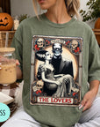 A woman wearing a green t-shirt with a graphic design featuring a couple and the text "MONSTER THE LOVERS". She is holding a drink in a glass.