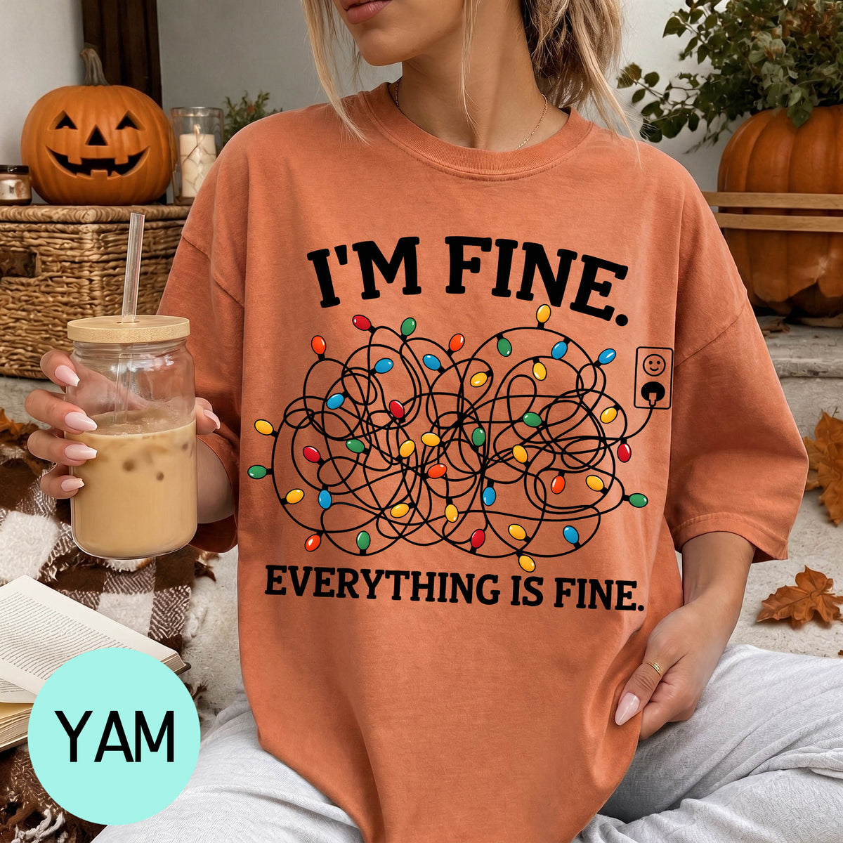 A woman wearing a t-shirt with a humorous graphic and text, sitting on a couch with a pumpkin and a mug of iced coffee in the background.