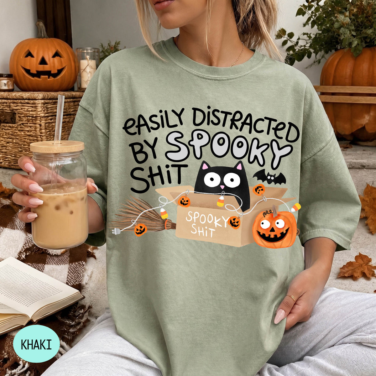 A woman wearing a green t-shirt with a humorous Halloween-themed graphic, including a cat, pumpkins, and bats, along with the text "EASILY DISTracted BY SPOKEY SHIT".