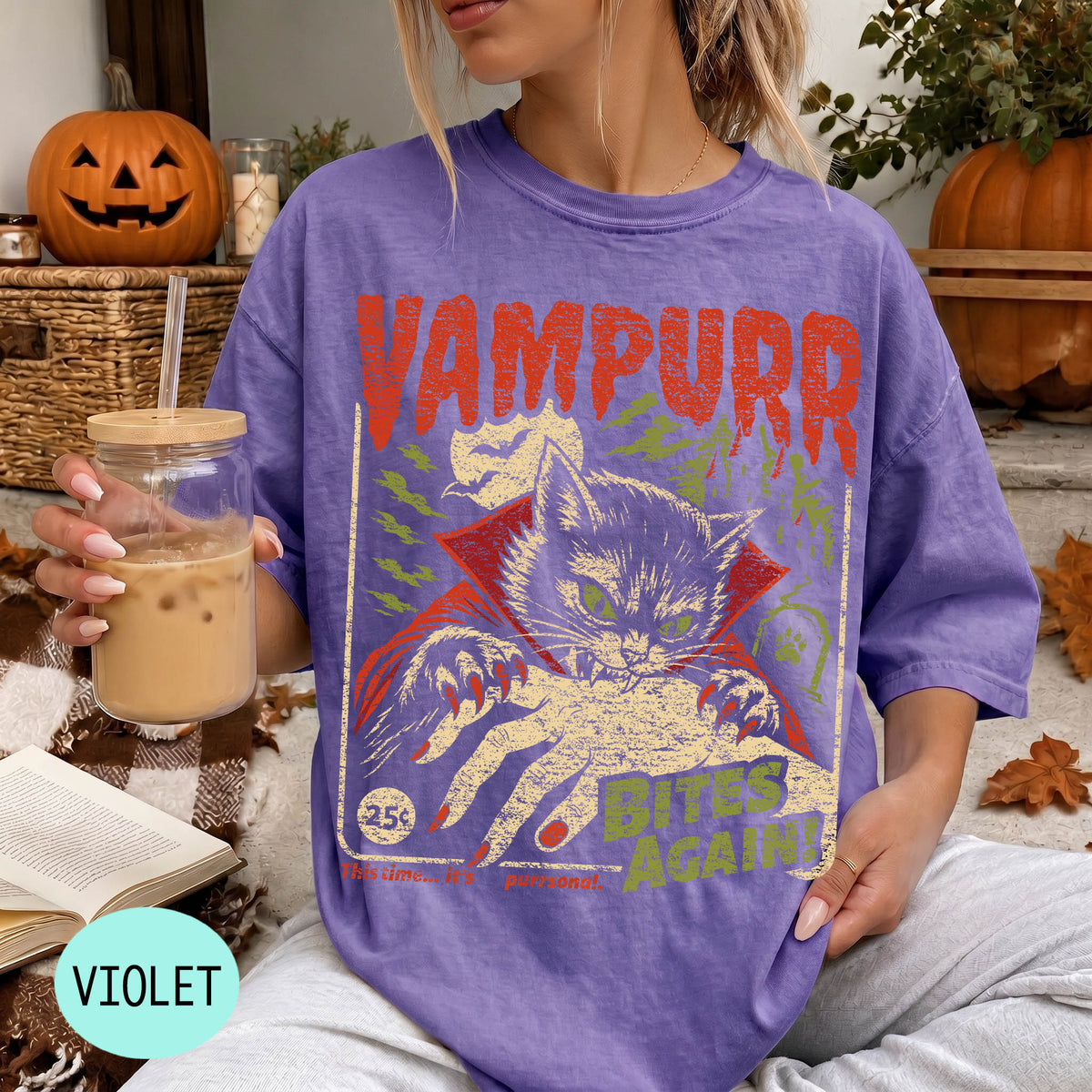 A woman wearing a purple t-shirt with a graphic design of a cat and the text "VAMPURR BITS AGAIN!" is sitting on a couch with a pumpkin and a book in the background.