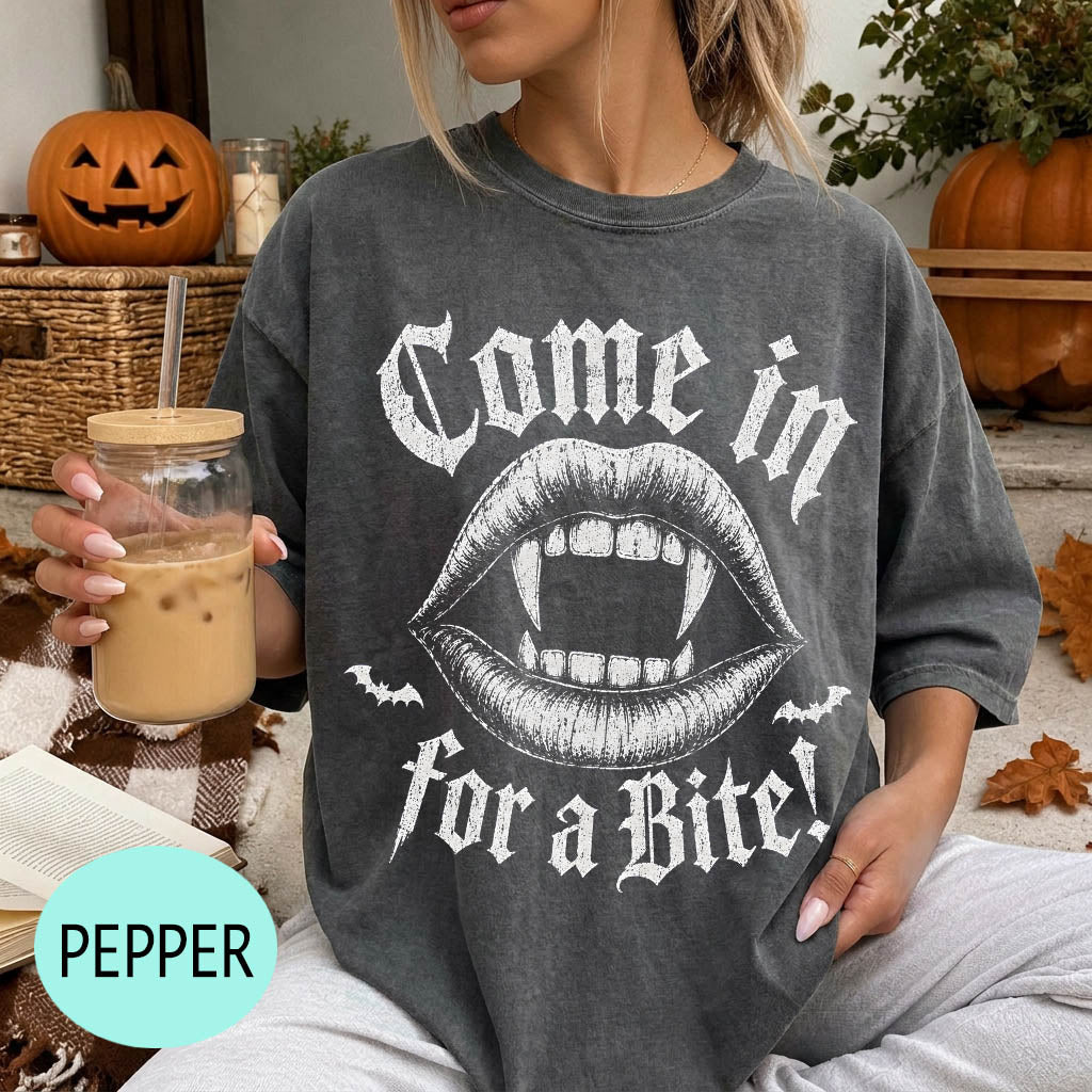 A woman wearing a t-shirt with a graphic design of a pair of open-mouthed bats and the text "Come in for a bite!" is sitting on a couch with a pumpkin and a mug of iced coffee in the background.