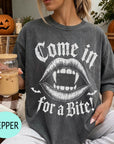 A woman wearing a t-shirt with a graphic design of a pair of open-mouthed bats and the text "Come in for a bite!" is sitting on a couch with a pumpkin and a mug of iced coffee in the background.