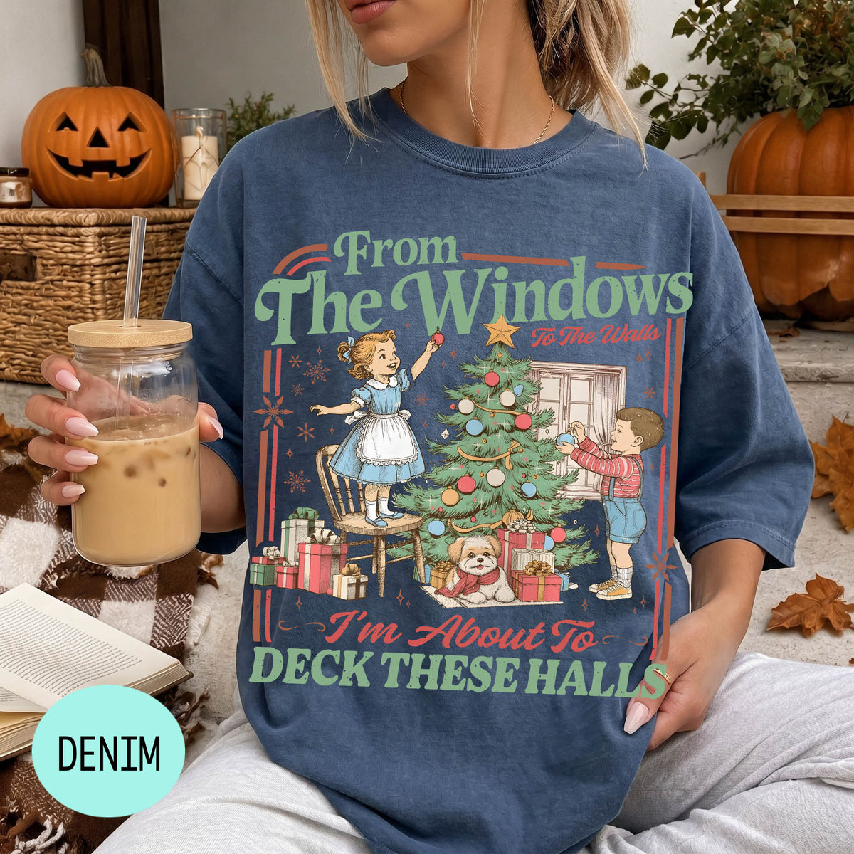 A woman wearing a blue t-shirt with a graphic design of a Christmas tree and two children decorating it, holding a drink in her hand.