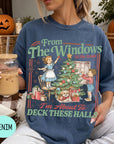 A woman wearing a blue t-shirt with a graphic design of a Christmas tree and two children decorating it, holding a drink in her hand.