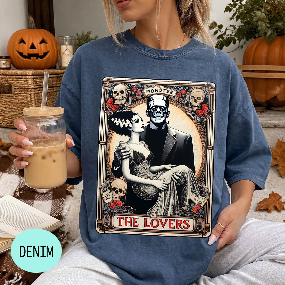 A woman wearing a blue t-shirt with a graphic design featuring a bride and groom, along with the text "MONSTER THE LOVERS". She is holding a drink in a glass and there are Halloween decorations visible in the background.