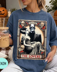 A woman wearing a blue t-shirt with a graphic design featuring a bride and groom, along with the text "MONSTER THE LOVERS". She is holding a drink in a glass and there are Halloween decorations visible in the background.