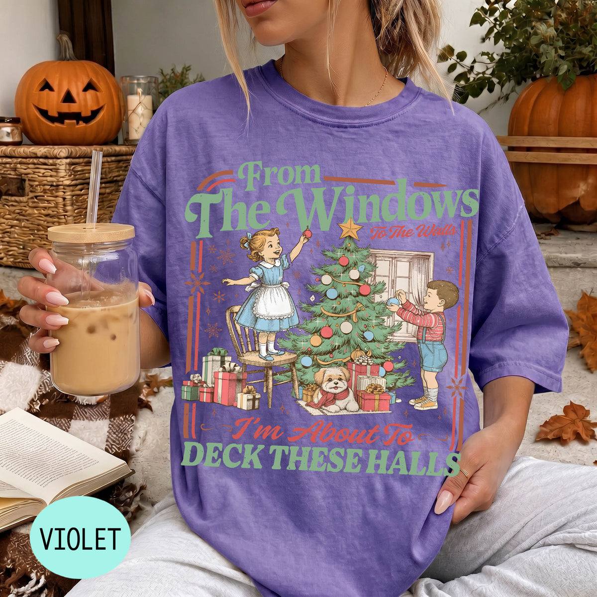 A woman wearing a purple t-shirt with a graphic design of a Christmas tree and two children decorating it, holding a drink in her hand.