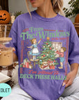 A woman wearing a purple t-shirt with a graphic design of a Christmas tree and two children decorating it, holding a drink in her hand.