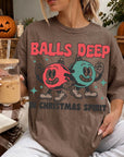 A woman wearing a t-shirt with a graphic design of two cartoon characters, one red and one blue, along with the text "BALLS DEEP IN CHRISTMAS SPIRIT". She is holding a glass of iced coffee and appears to be sitting on a couch or chair.