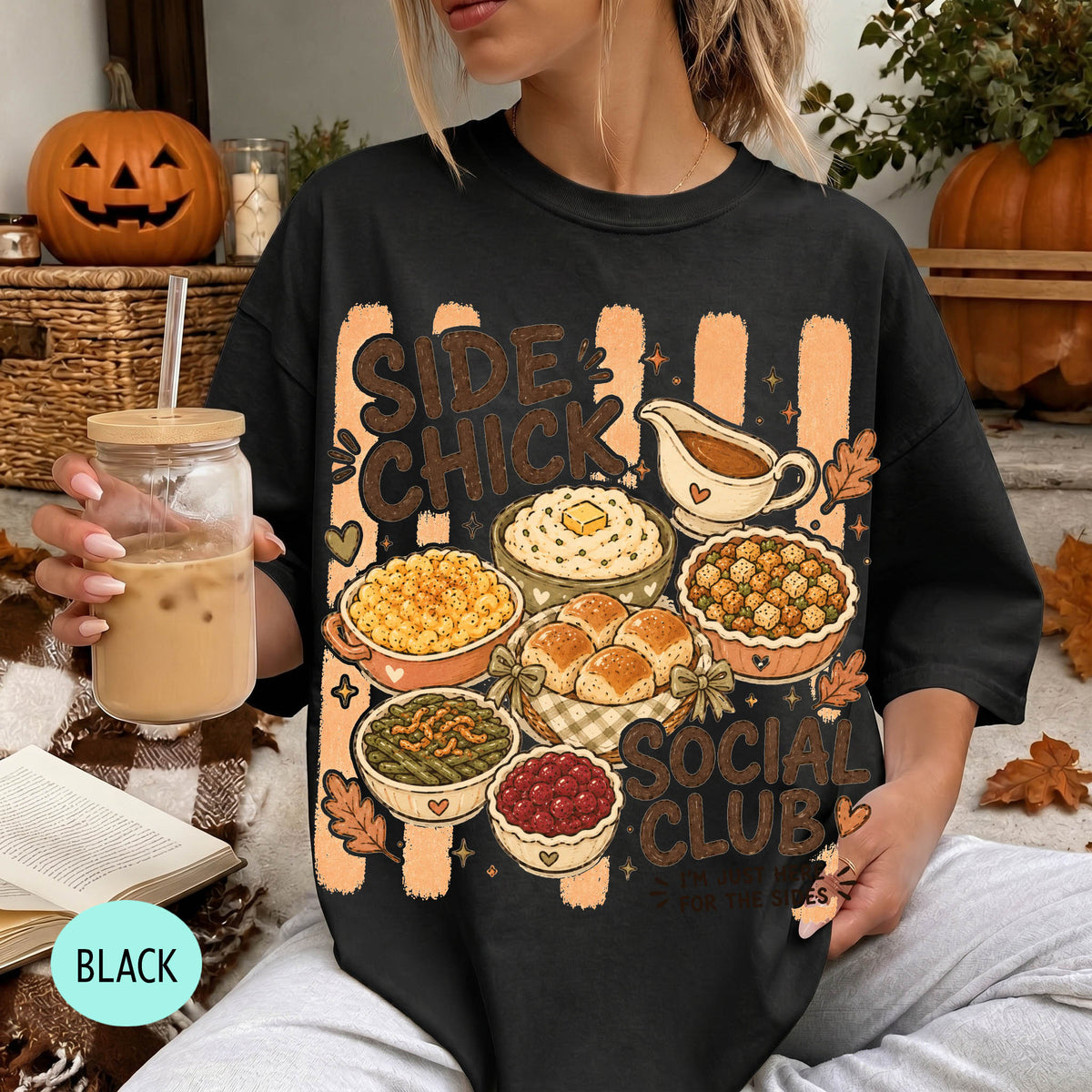 A woman wearing a black t-shirt with a graphic design of various Thanksgiving dishes, including a pumpkin pie, mashed potatoes, stuffing, and cranberry sauce. She is holding a drink in a glass with a straw.