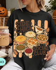 A woman wearing a black t-shirt with a graphic design of various Thanksgiving dishes, including a pumpkin pie, mashed potatoes, stuffing, and cranberry sauce. She is holding a drink in a glass with a straw.