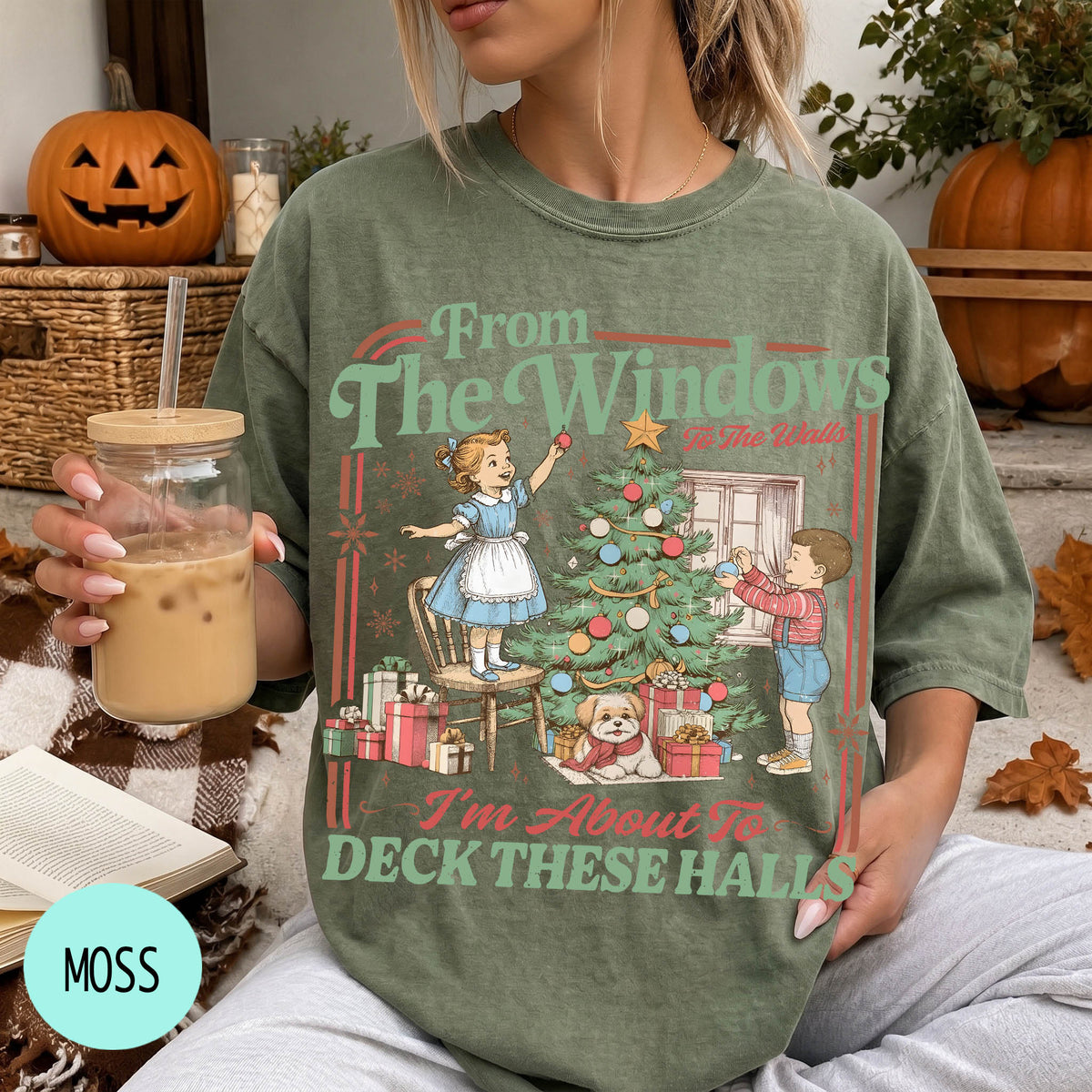 A woman wearing a green t-shirt with a Christmas-themed illustration and text, holding a glass of iced coffee.