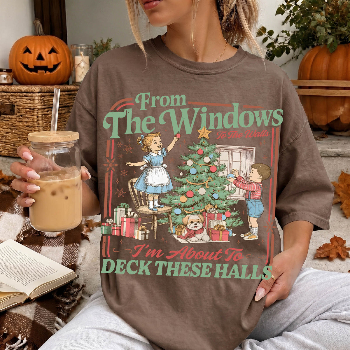 A woman wearing a t-shirt with a graphic design of a Christmas tree and two children decorating it, holding a drink and sitting on a couch with a book and a pumpkin decoration in the background.
