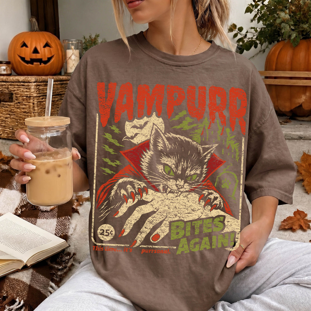 A woman wearing a t-shirt with a graphic design of a cat and the text "VAMPURR BITE'S AGAIN" is sitting on a couch with a pumpkin and a drink in her hand.