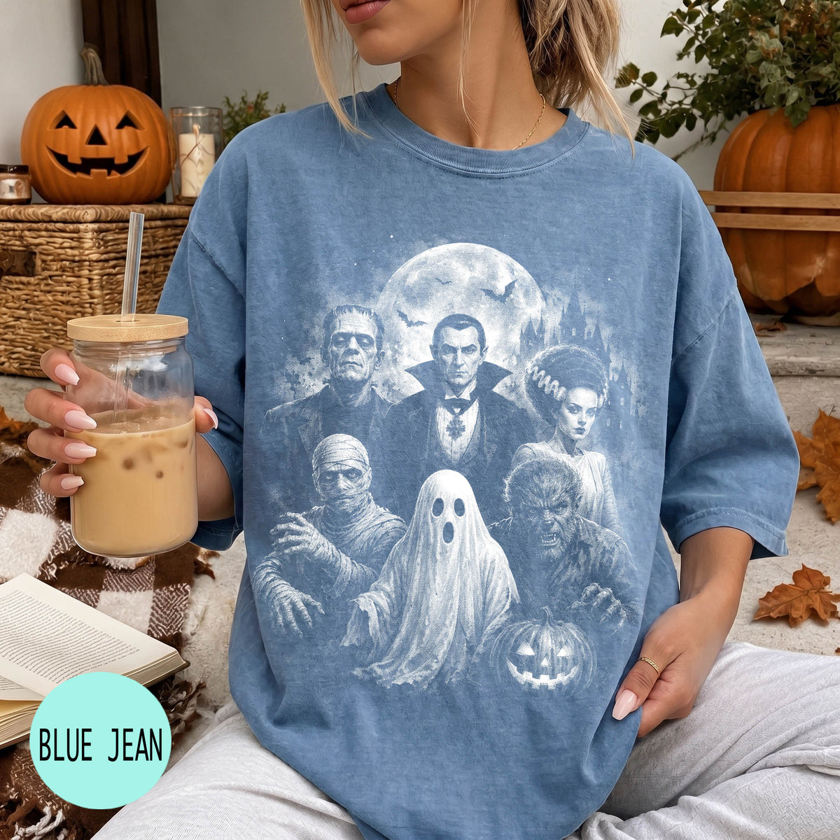 A woman wearing a blue t-shirt with a Halloween-themed graphic design, holding a drink and sitting on a couch with a pumpkin and other Halloween decorations in the background.
