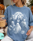 A woman wearing a blue t-shirt with a Halloween-themed graphic design, holding a drink and sitting on a couch with a pumpkin and other Halloween decorations in the background.