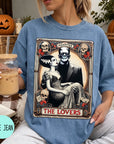 A woman wearing a blue t-shirt with a graphic design featuring a couple and the text "MONSTER THE LOVERS". She is holding a drink in a glass with a straw.